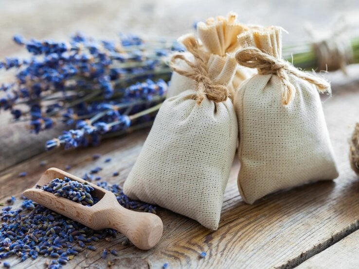 Drei mit Kordel verschlossene Duftsäckchen und ein Holzlöffel liegen auf einem hölzernen Tisch neben Lavendel.