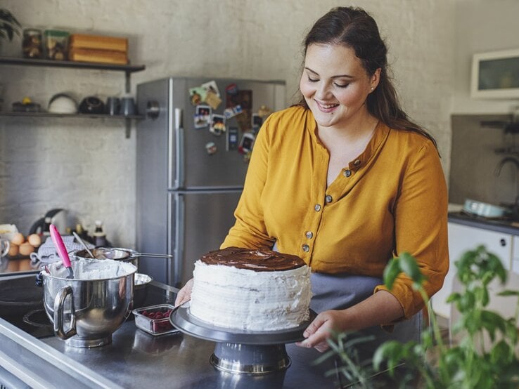 Junge Frau mit braunen Haaren und gelber Bluse dekoriert in einer modernen Küche lächelnd eine selbst gemachte Sahnetorte.