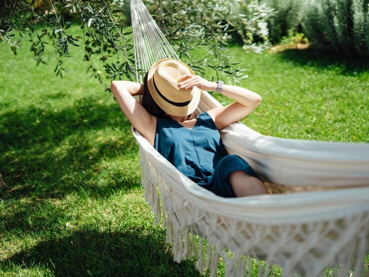 Eine Frau in blauem Kleid liegt entspannt mit Strohhut in einer Hängematte auf einer sonnigen Wiese.
