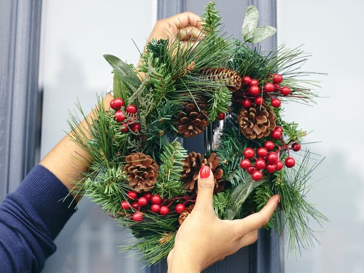 Hände einer Person mit rotem Nagellack hängen einen Weihnachtskranz aus Tannenzweigen, Zapfen und roten Beeren an eine blaue Tür.
