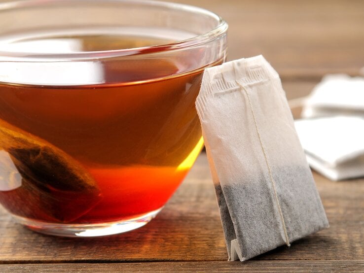 Ein gefülltes Glas Tee mit einem eintauchenden Teebeutel steht auf einem Holztisch, daneben ein unbenutzter Teebeutel. | © Adobe Stock/MK studio