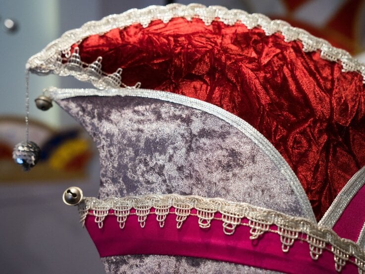 Nahaufnahme einer detailreichen Narrenkappe aus rotem Samt und grauem Stoff, verziert mit goldener Spitze und kleinen Glöckchen.