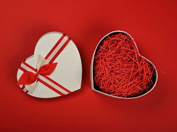 Geöffnete herzförmige Geschenkbox mit rotem Papierstreifen-Füllmaterial auf rotem Hintergrund, der Deckel mit Schleife liegt daneben.