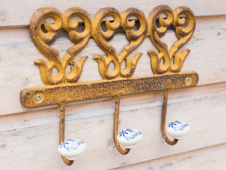 Drei antike, goldgelbe Haken mit filigranen Verzierungen und weißen Keramikknäufen mit blauen Blumenmotiven an einer Holzlattenwand.