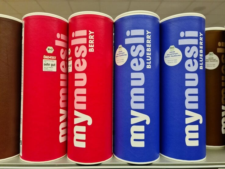 Vier Müslidosen der Marke mymuesli, zwei in Rot für Beerenmüsli und zwei in Blau für Blaubeermüsli, stehen nebeneinander im Regal.