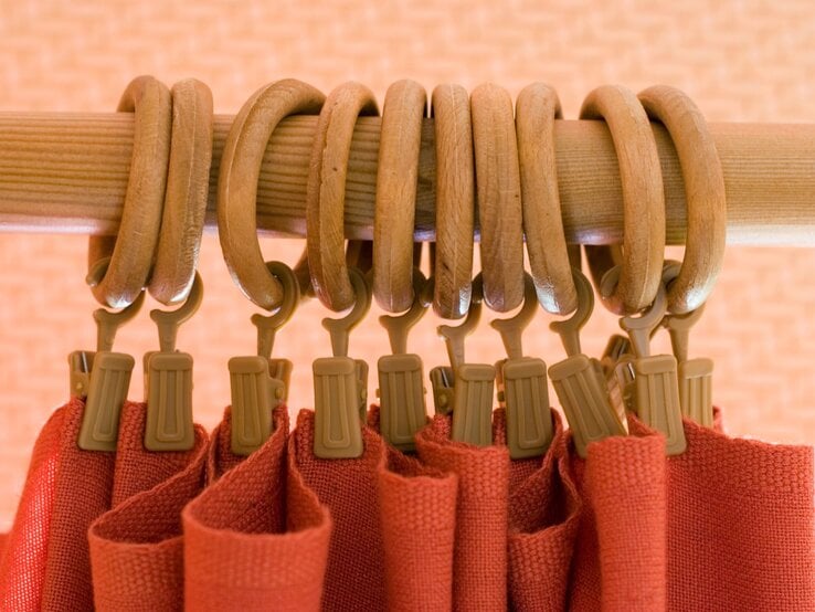 Nahaufnahme einer Holzgardinenstange mit mehreren Gardinenringen aus Holz und beigefarbenen Clips, an denen ein roter Stoffvorhang befestigt ist.