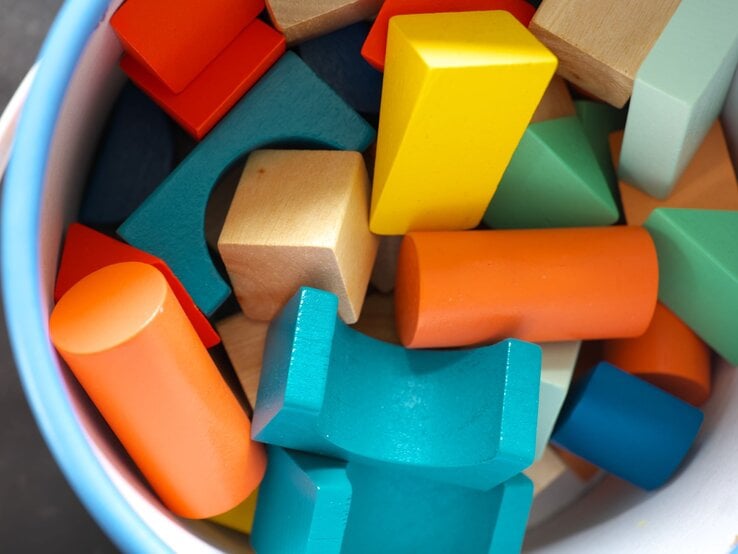 Ein Eimer voller bunter Holzbausteine in verschiedenen Formen, darunter Zylinder, Würfel und Dreiecke in Blau, Orange, Gelb und Grün.