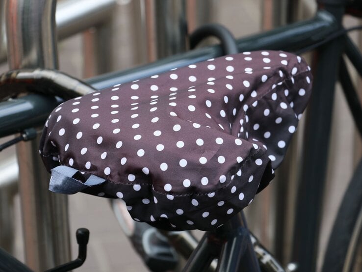 Nahaufnahme eines Fahrradsattels mit wasserfestem, braunem Überzug mit weißen Punkten, befestigt an einem geparkten Fahrrad vor Metallstangen.