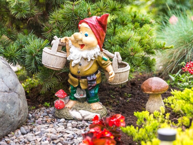 Ein bunter Gartenzwerg mit roter Mütze und weißen Bart trägt zwei Holzeimer und steht lachend zwischen grünen Pflanzen und Pilzen.
