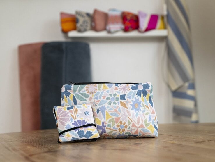 Zwei Kosmetiktaschen mit floralen Mustern in Pastellfarben stehen auf einem Holztisch, im Hintergrund Stoffe und Deko.
