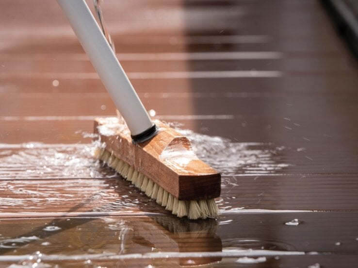 Grobe Bürste mit Holzgriff schrubbt unter fließendem Wasser eine nasse, braune Terrassendiele bei Sonnenlicht.