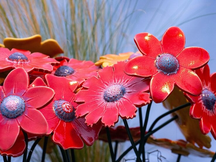 Leuchtend rote DIY-Gartenstecker mit blauen Mitteln glänzen im Sonnenlicht vor unscharfem Gras und blassem Himmel.