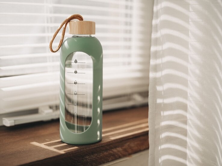 Moderne Glasflasche mit DIY-Wasser-Tracker leuchtet im warmen Licht vor halb geöffneten Jalousien.