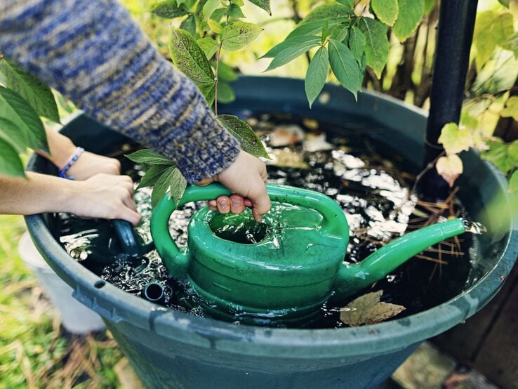 Zu sehen ist eine Regentonne, die mit Wasser gefüllt ist und Hände, die mit einer Gießkanne das Wasser herausschöpfen