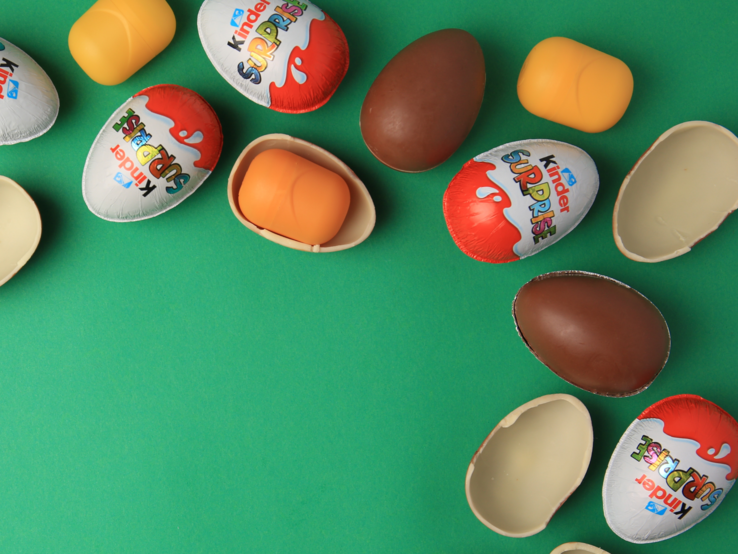 Mehrere bunte Überraschungseier, teils geöffnet mit gelben Kapseln, liegen verstreut auf grünem Hintergrund.
