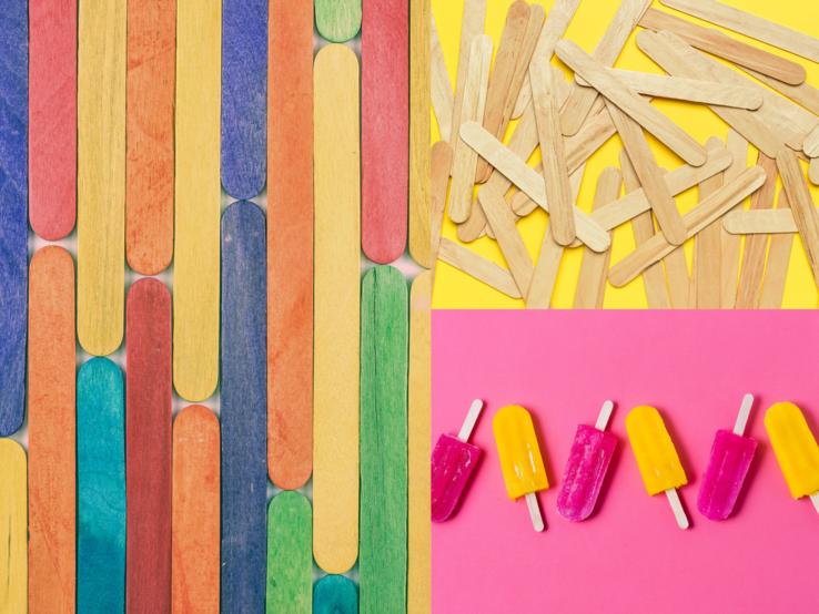 Bunte Eisstiele liegen geordnet, lose verstreut und als pink-gelbe Eis am Stiel auf knallig pink-gelb geteiltem Hintergrund.