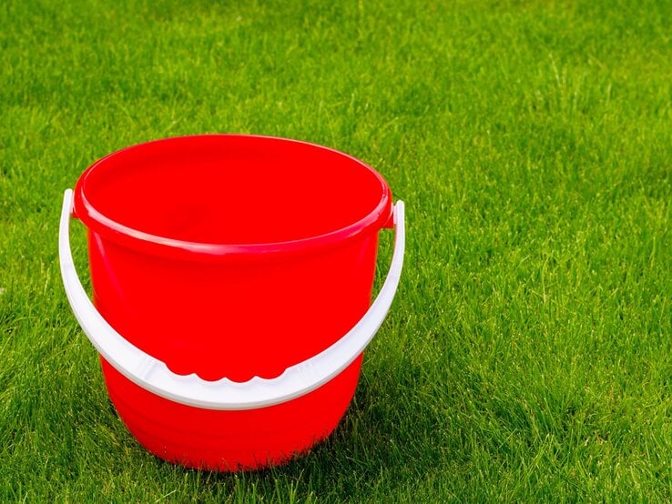Ein leerer, knallroter Plastikeimer mit weißem Henkel steht auf dichtem, frisch gemähtem, sattem grünen Rasen.