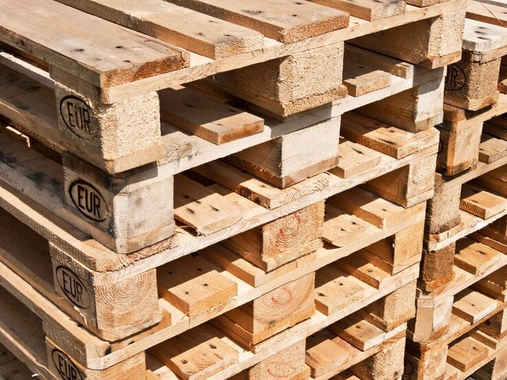 Stapel aus hellem Holz gefertigten Europaletten mit sichtbarem EUR-Logo, teils mit Gebrauchsspuren, in warmem Sonnenlicht aufgenommen.