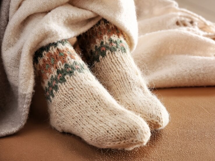 Kuschelige gestrickte Wollsocken mit farbigem Norwegermuster ragen aus beiger Decke, liegend auf einem weichen Sofa in warmem Licht.