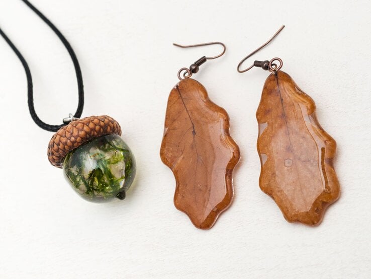 Handgefertigte Eichel-Halskette mit grünem Glasanhänger und zwei glänzenden Ohrringen in Form brauner Eichenblätter auf Weiß.
