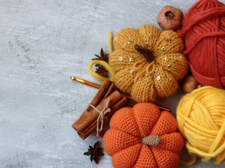 Herbstliche Deko mit orangefarbenen Häkelkürbissen, glänzender Wolle, Zimt, Sternanis und goldener Häkelnadel auf Betonhintergrund.