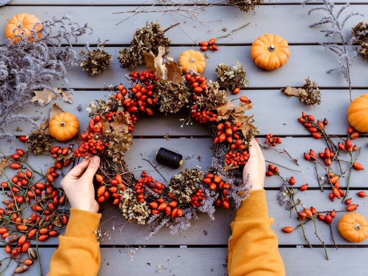 Zwei Hände in senfgelbem Pullover halten einen herbstlichen Kranz aus Hagebutten, Trockenblüten und Eichenblättern auf Holztisch.