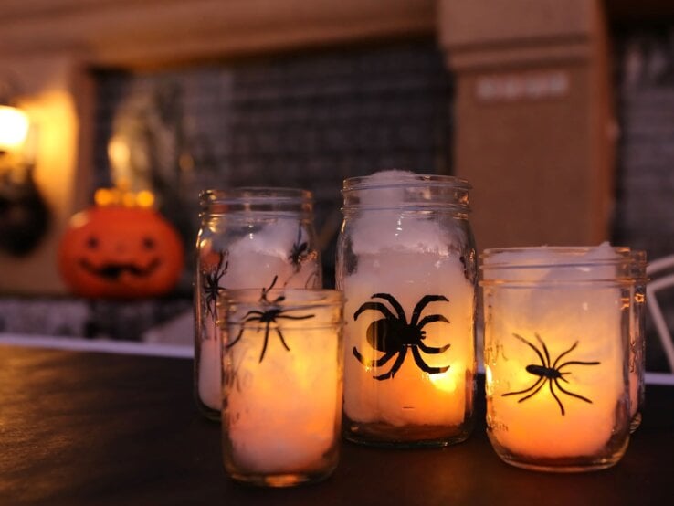 Vier Einmachgläser mit leuchtenden Kerzen und Spinnenmotiven stehen auf einem Tisch vor verschwommenem Halloween-Dekor.