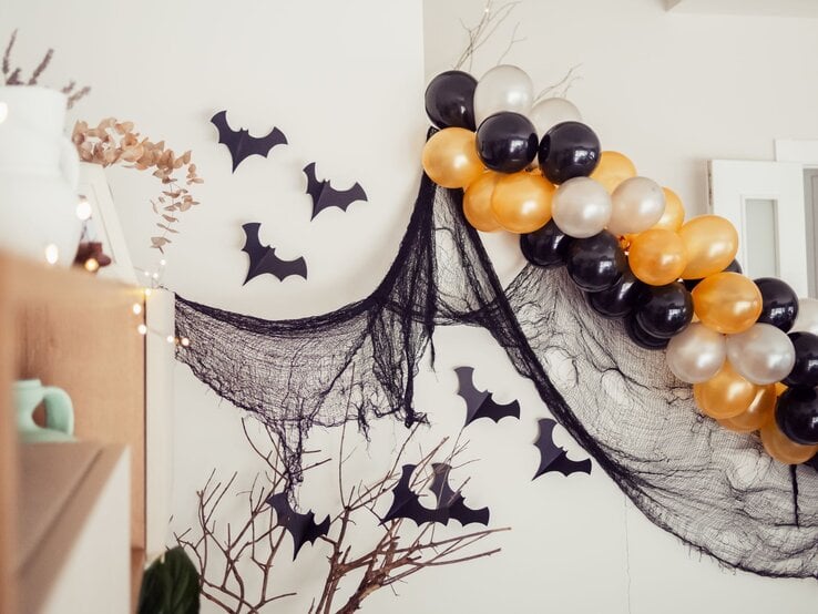 Halloween-Deko mit schwarzen Stoffnetzen, Papierfledermäusen und glänzenden Luftballons in Schwarz, Gold und Perlweiß.