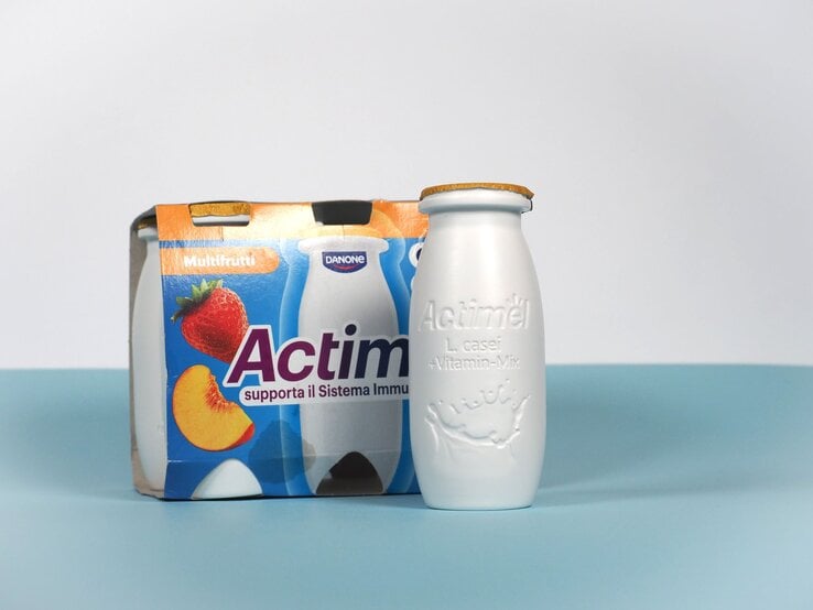 Eine weiße Actimel-Flasche mit goldener Versiegelung steht vor einem bunten Viererpack in Multifrucht-Design auf blauem Untergrund.