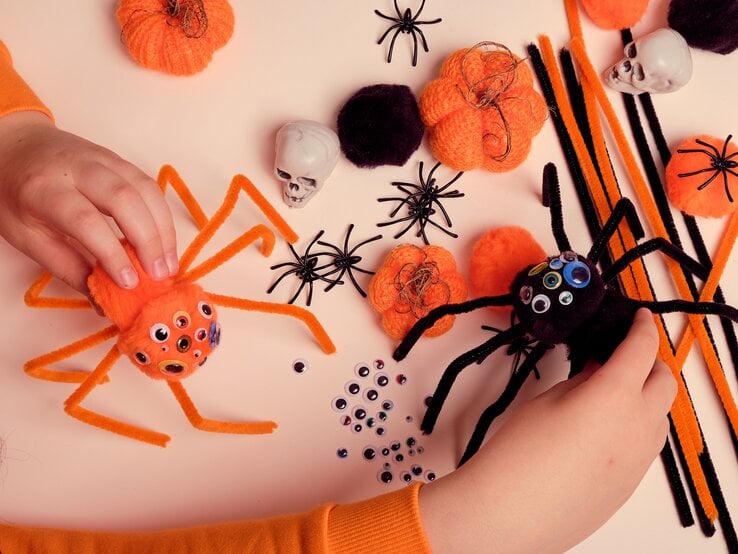 Nahaufnahme von zwei Filzspinnen mit Pfeifenreinigern, Dekokürbisse, Schädel und viele Wackelaugen liegen verstreut auf hellbeigem Basteltisch.