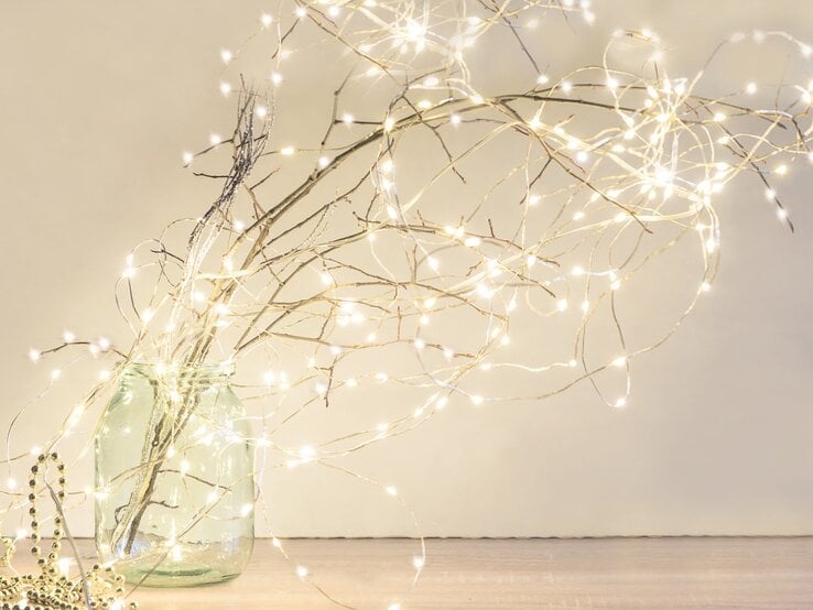 Ein bulliges glas mit Zweigen, die mit Lichterketten geschmückt sind, Hintergrund hell und die Lichterketten leuchten goldenweiss