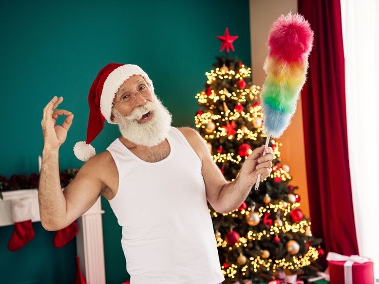 Basteln mit Staubwedel: Älterer Mann mit weißem Rauschebart und Santa-Mütze posiert lachend mit Staubwedel vor festlich geschmücktem Weihnachtsbaum.
