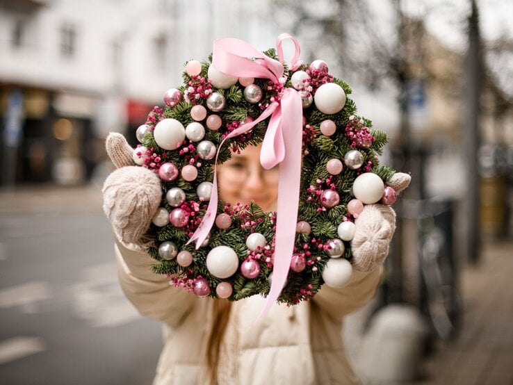 Junge Frau in Winterjacke und Wollhandschuhen hält draußen einen üppigen Adventskranz mit rosa Kugeln und Schleife.