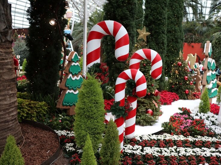 Riesige rot-weiße Zuckerstangen stehen zwischen Weihnachtsbäumen, Blumenbeeten und Lebkuchenfiguren in festlicher Kulisse.