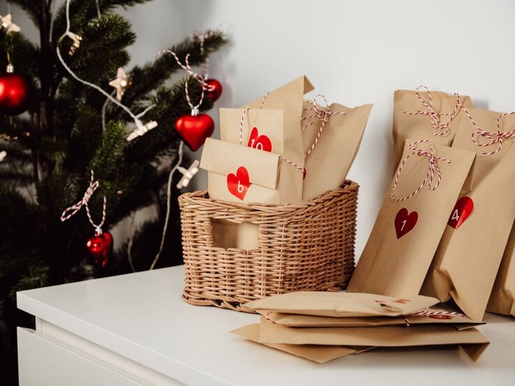 Liebevoll verpackte Adventskalendertüten mit roten Herzen und Zahlen liegen neben einem geschmückten Weihnachtsbaum auf Weiß.