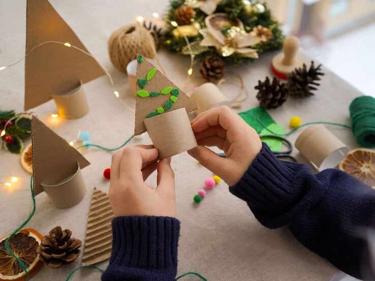 Weihnachtliche Teelichthalter mit Zapfen, Filz, Lichterkette und Trockenobst, Kind hält fertigen Baum aus Klopapierrollen in den Händen.