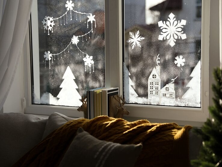 Winterliche Fensterdeko mit Schneeflocken, Tannen und Häuschen, davor Sofa mit Kissen und gelber Strickdecke.