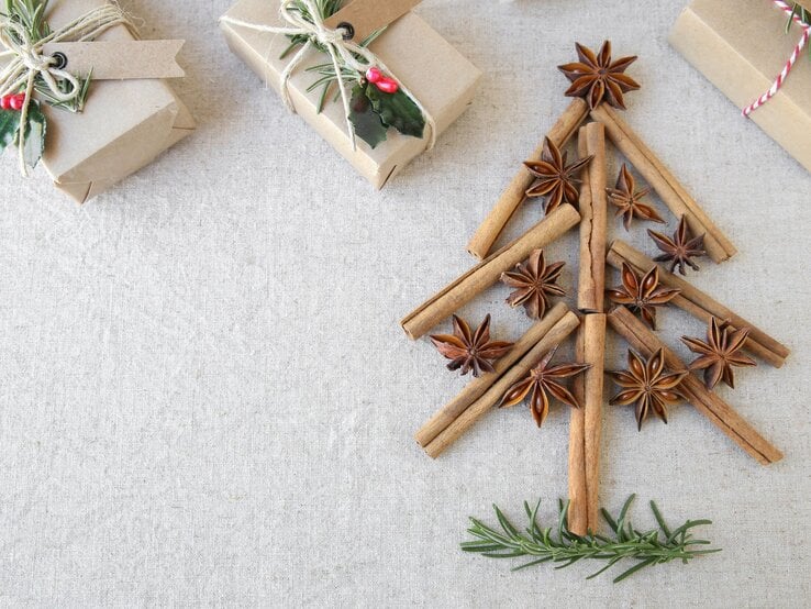 Aus Zimtstangen, Sternanis und Rosmarin gelegener Weihnachtsbaum auf Leinentuch, daneben schlicht verpackte Geschenke.