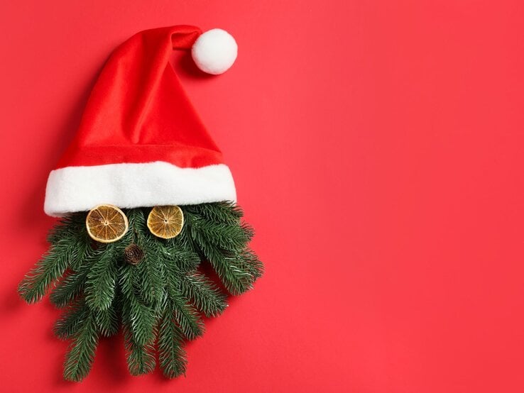 Tannenzweige mit getrockneten Orangenscheiben und Zapfen als Gesicht unter roter Weihnachtsmütze auf rotem Hintergrund.