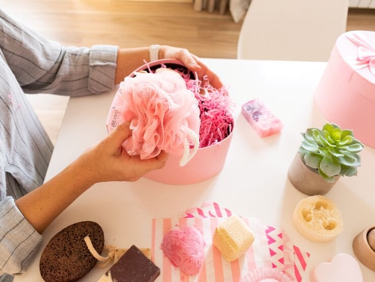 Frau in gestreifter Bluse hält rosa Duschschwamm über geöffneter Geschenkbox mit Pflegeprodukten auf hellem Tisch.