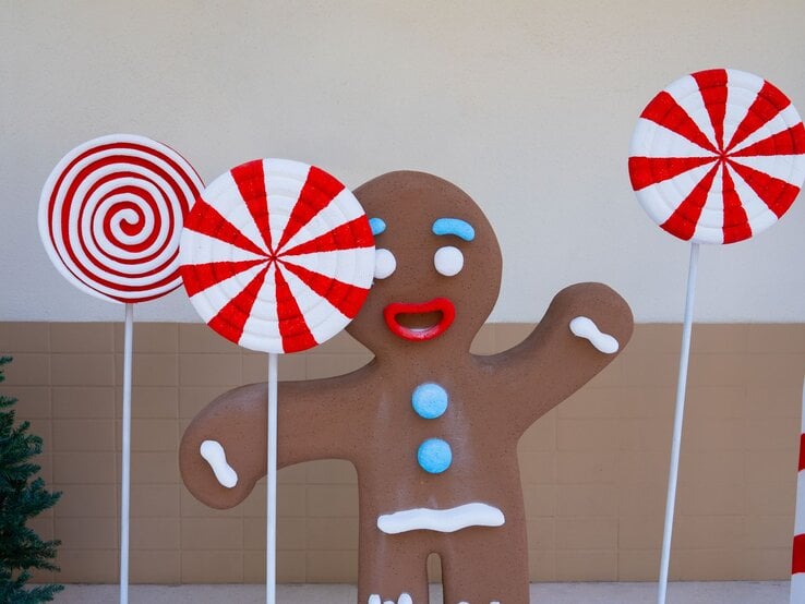 Fröhliche Lebkuchenfigur mit roten Lippen und blauen Knöpfen steht zwischen riesigen rot-weißen Zuckerstangen-Lollis.