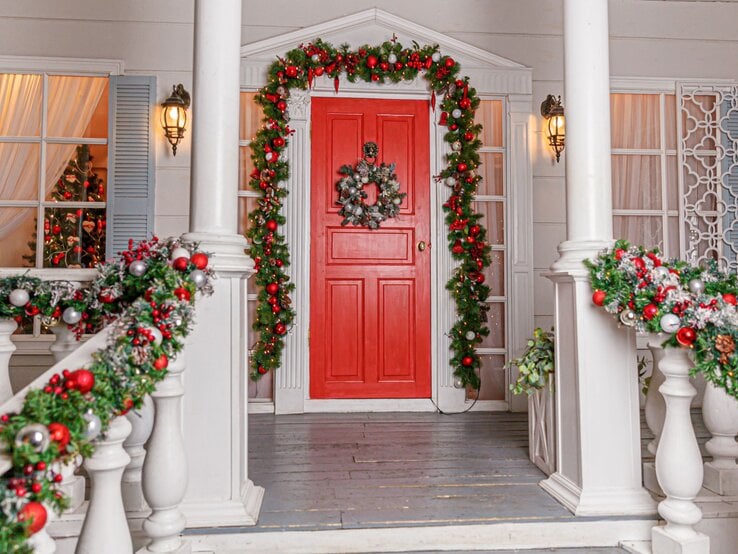 Weihnachtlich geschmückte Veranda mit roter Tür, grünen Girlanden und silbernem Kranz, im Fenster leuchtet ein Christbaum.