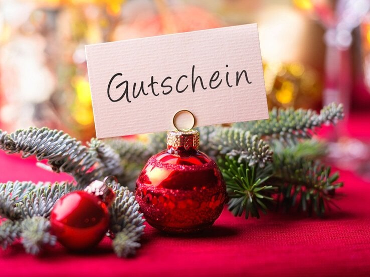 Rote Christbaumkugel mit Kärtchenaufsatz „Gutschein“ liegt auf Tannenzweig vor festlich verschwommenem Hintergrund.