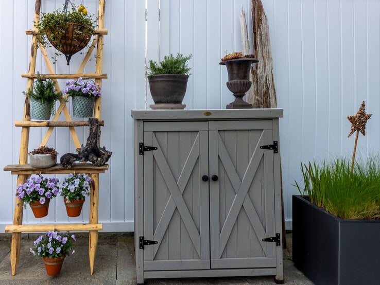 Blühende violette Blumen, Tontöpfe und Kräuter arrangiert auf Leiterregal neben rustikalem Gartenschrank aus Holz.