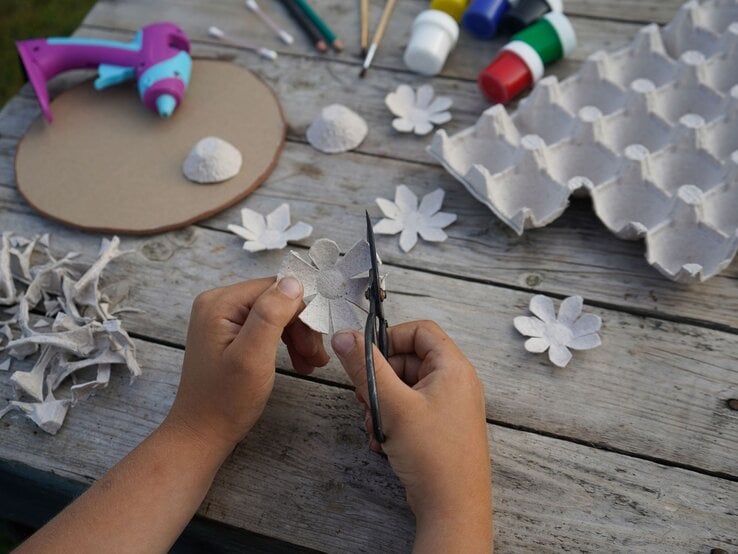 Kinderhände schneiden aus grauer Eierkartonpappe eine Blume, umgeben von Bastelmaterialien auf einem Holztisch.