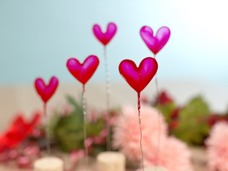 Herzförmige Deko auf Draht vor pastellblauem Hintergrund und verschwommenem Blumenarrangement aus Rosa und Rot.