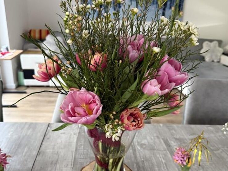 Ein bunter Frühlingsstrauß mit rosa Tulpen und kleinen weißen Blüten steht in einer Vase auf grauem Holztisch.