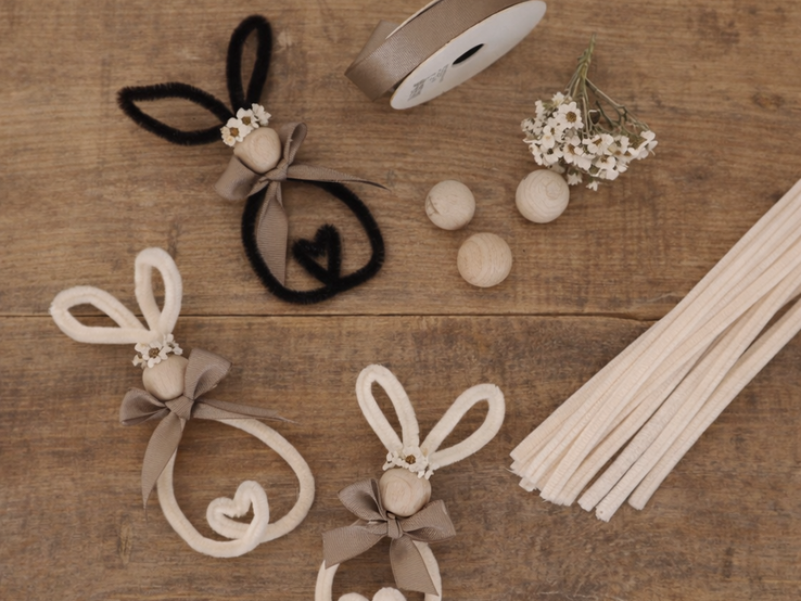 Drei kleine Osterhasen aus Pfeifenreiniger und Holzperlen liegen auf rustikalem Holztisch, daneben Bandrolle, Blütenzweig und Holzstäbe.