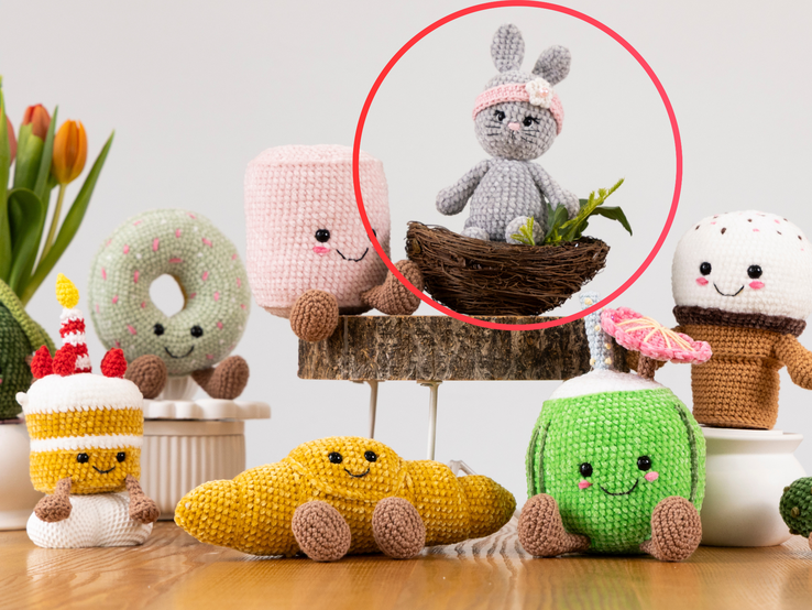 Ein gehäkelter grauer Hase mit rosa Stirnband und Blüte sitzt in einem kleinen Vogelnest, umgeben von grünen Pflanzen auf einem Holzpodest.