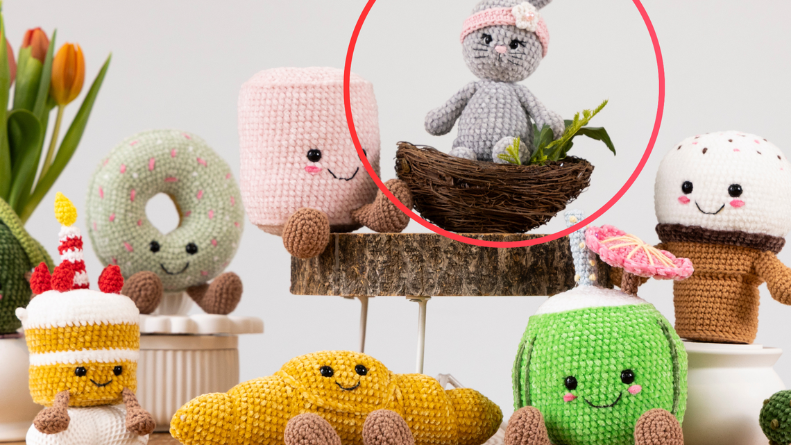 Ein gehäkelter grauer Hase mit rosa Stirnband und Blüte sitzt in einem kleinen Vogelnest, umgeben von grünen Pflanzen auf einem Holzpodest.